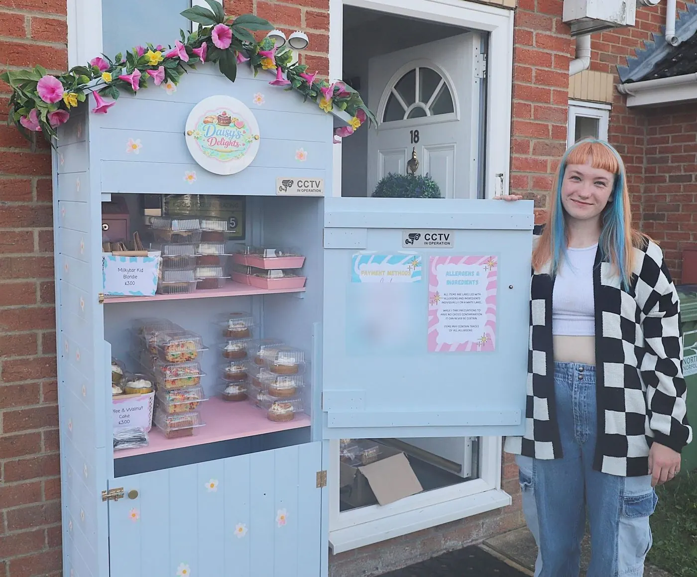 Daisy standing beside the pale blue Daisy's Delights bake shed, its shelves full of boxed slices and cupcakes